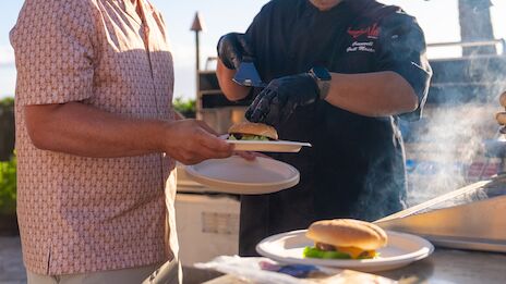 Two people grilling burgers at an outdoor barbecue, with one serving a burger onto a plate as another cooks, steam rising from the grill.