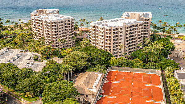 Aerial view of a coastal hotel complex with high-rise buildings, palm trees, and bright orange tennis courts near the ocean, surrounded by greenery.