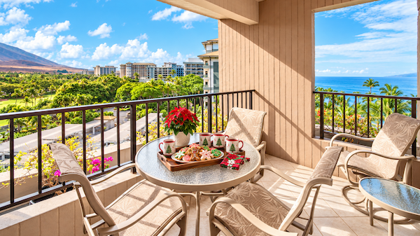 A sunny balcony with a round table and four cushioned chairs overlooks a tropical ocean view, with a plate of snacks and flowers on the table.