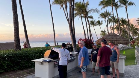 People grilling by a roadside barbecue near a beach, with tall palm trees and a sunset over the ocean. Ends with a period.