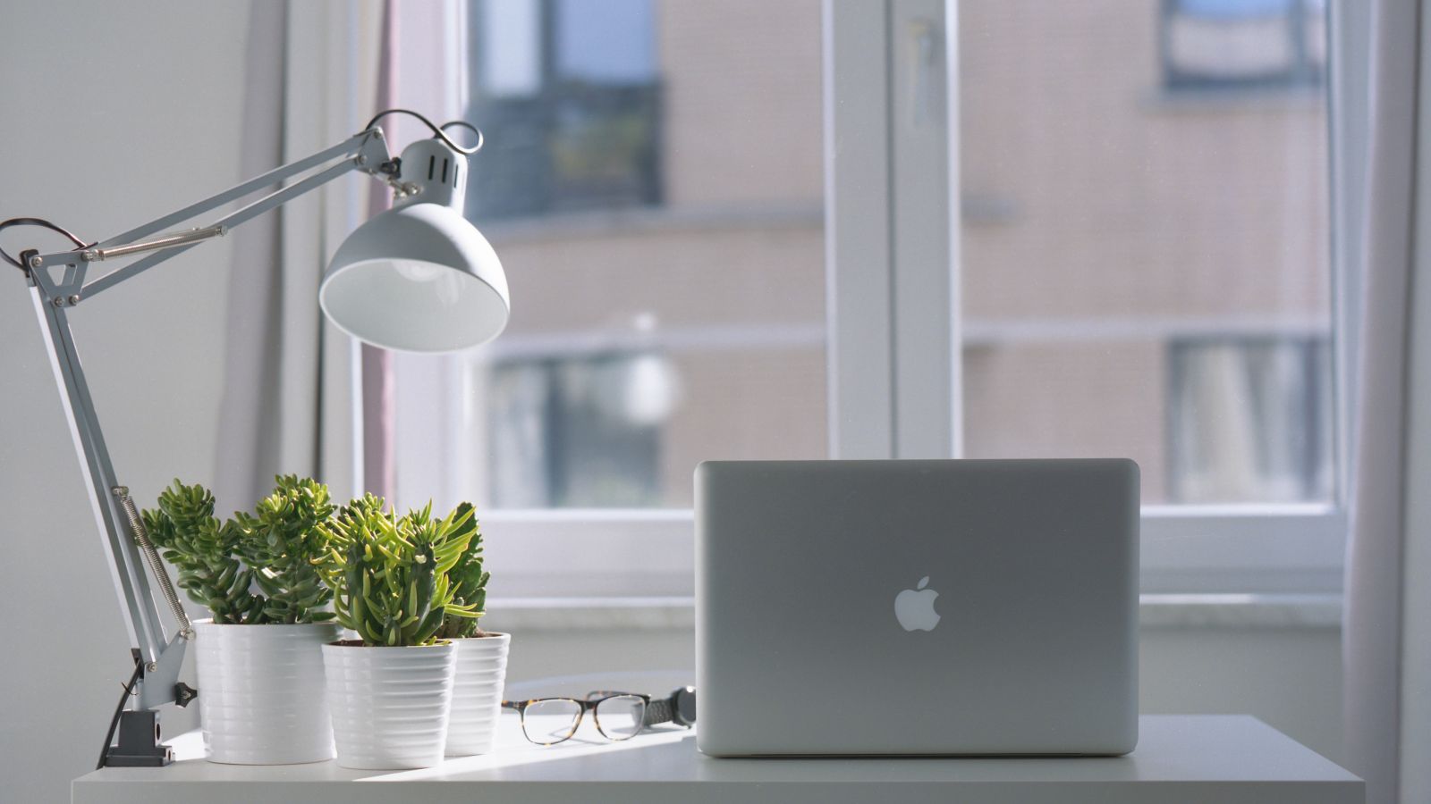 A minimalist desk setup with a lamp, potted plants, a laptop, and a pair of glasses, situated by a window.