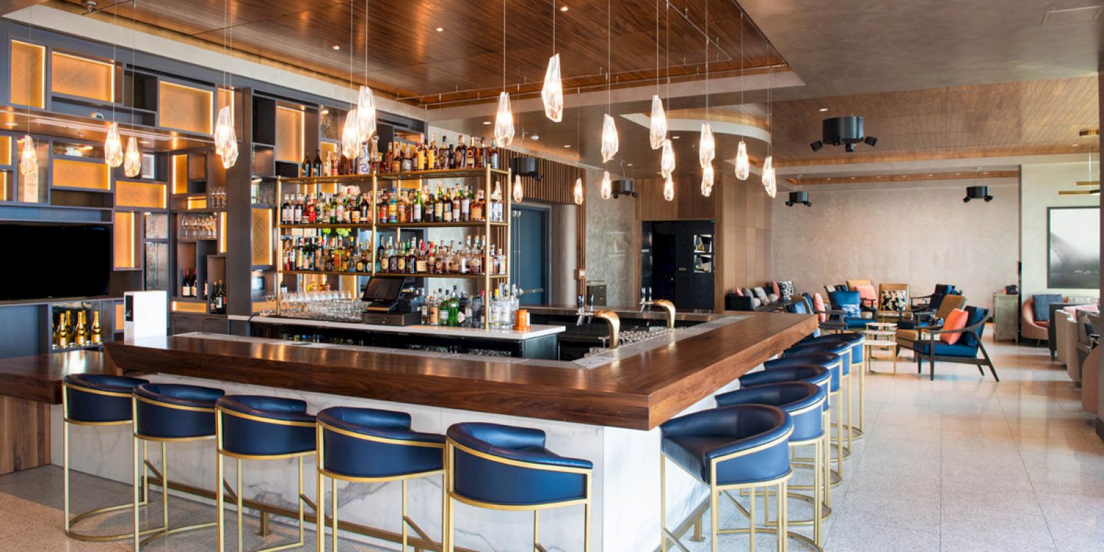 This image depicts a modern bar with wooden accents, a well-stocked bar, blue barstools with gold frames, and cozy seating areas in the background.