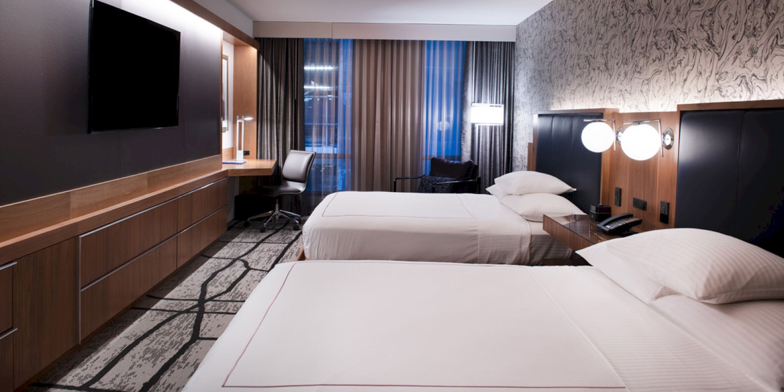 A modern hotel room with two double beds, wall-mounted TV, a desk, a chair, and stylish decor including patterned wallpaper and floor-to-ceiling curtains.