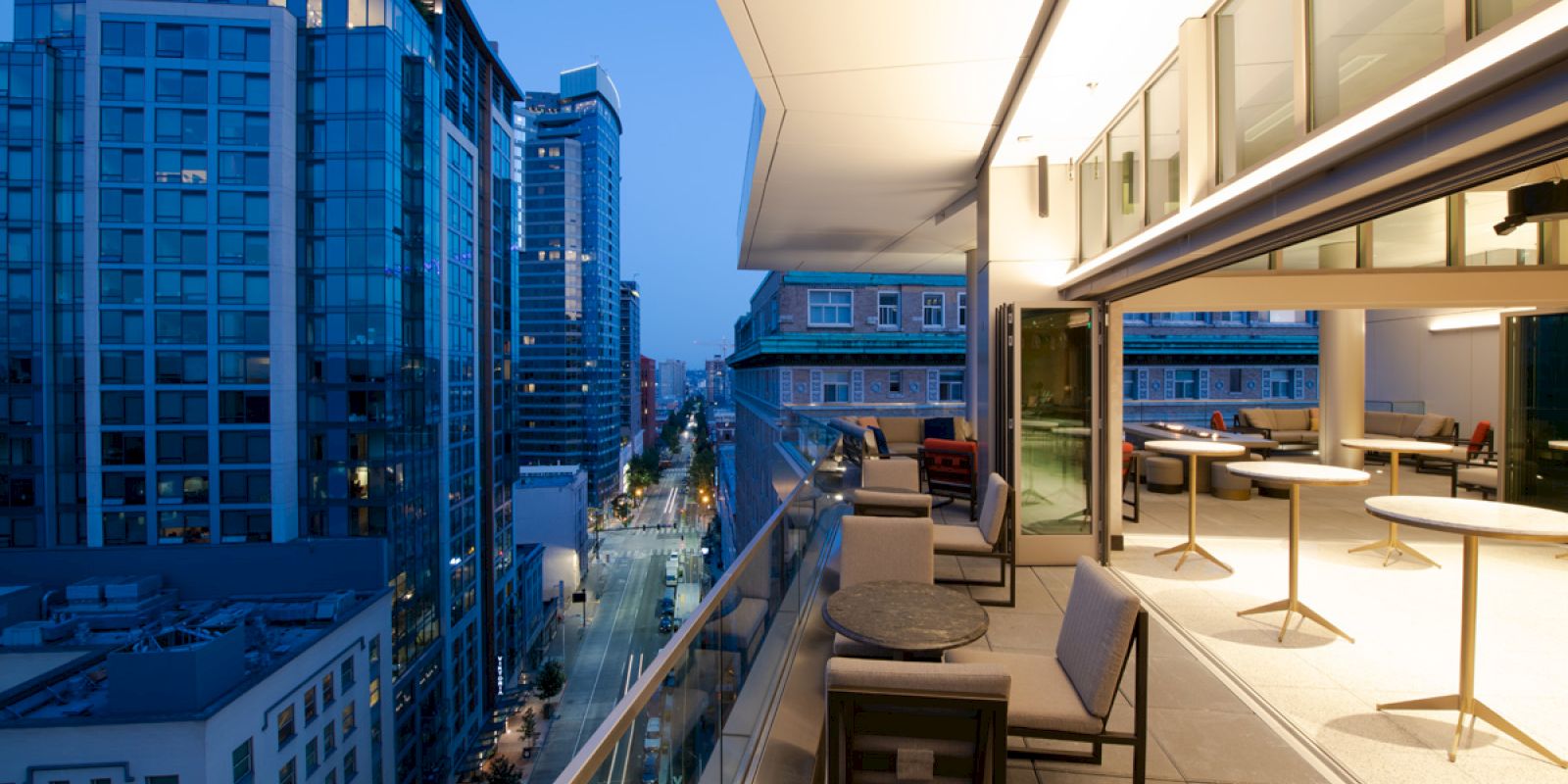 A modern balcony with furniture overlooks a cityscape of tall buildings at dusk. The interior of the adjacent building is well-lit.