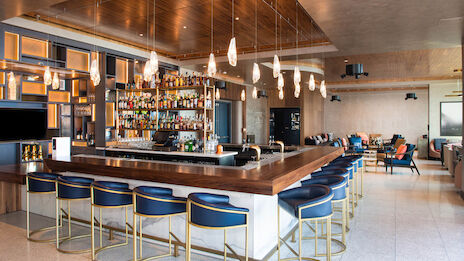 A modern bar with stylish seating, a well-stocked liquor shelf, hanging lights, and a lounge area in the background.