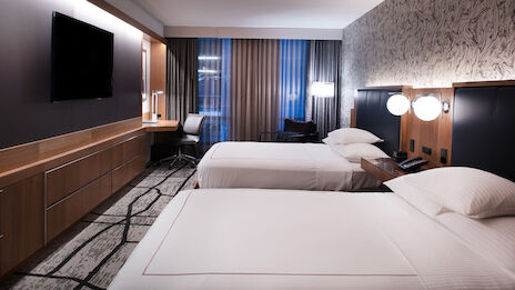 A modern hotel room with two beds, a flat-screen TV, a desk and chair, contemporary decor, and large windows with curtains ending the sentence.