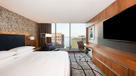 A modern hotel room with a large bed, wall-mounted TV, desk, and chair. Large windows provide a view of surrounding buildings and water.