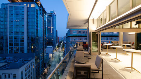 A modern rooftop terrace with outdoor seating overlooks a cityscape with tall buildings at dusk, showcasing a mix of urban and relaxed ambience.