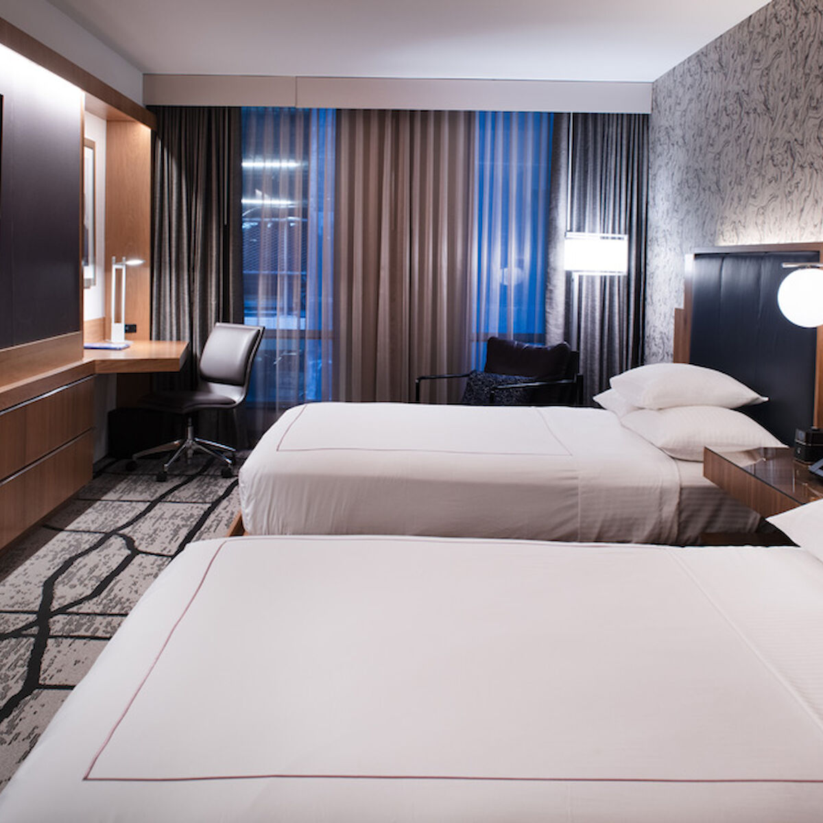 This image shows a modern hotel room with two beds, a wall-mounted TV, a work desk, chairs, bedside lamps, and stylish decor.