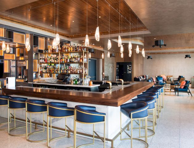 The image shows a modern bar with a wooden counter, blue and gold stools, hanging lights, and a well-stocked bar area. It has a cozy seating space.