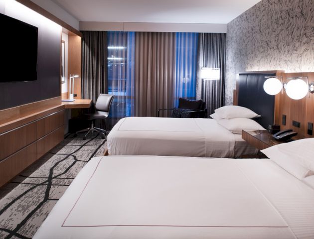 The image shows a modern hotel room with two neatly made beds, a large TV, a work desk by the window, and stylish lighting fixtures.