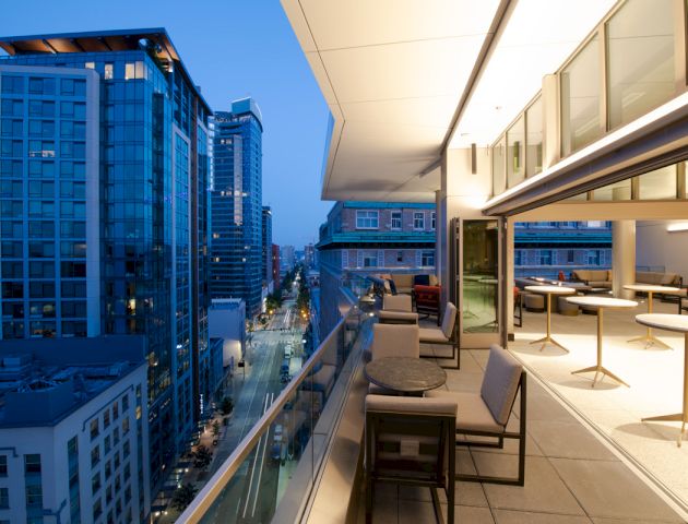 A high-rise outdoor patio with tables and chairs overlooks a city skyline at dusk, featuring modern buildings and illuminated windows.