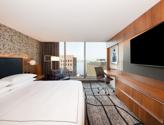 A modern hotel room with a large bed, TV, desk, chair, and a window with a city view. The design features wooden accents and a patterned wall.