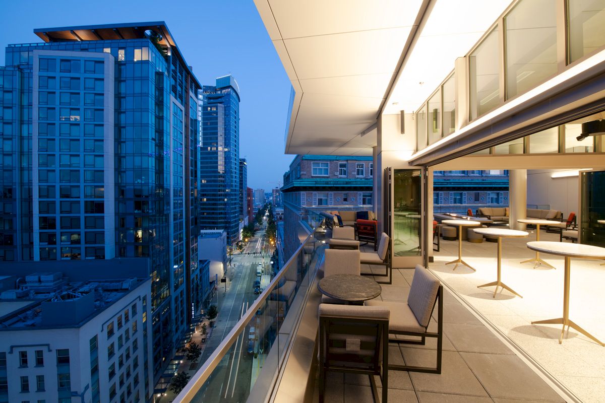 A modern urban balcony with a seating area overlooks a city street lined with high-rise buildings, under dusk lighting.