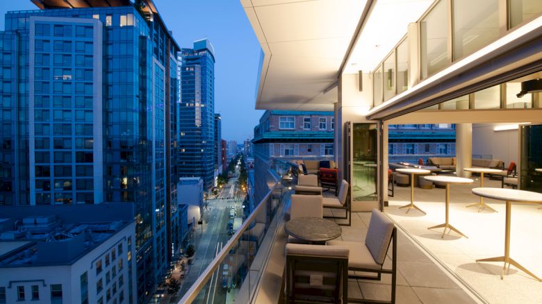 A modern urban balcony with a seating area overlooks a city street lined with high-rise buildings, under dusk lighting.