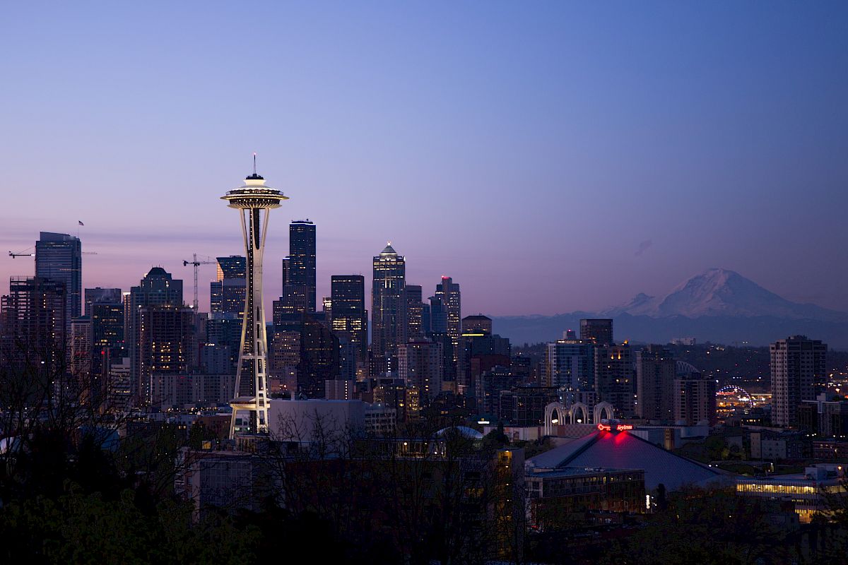 The image features a city skyline at dusk with a prominent tower in the foreground and a mountain in the background. The city lights are starting to glow.