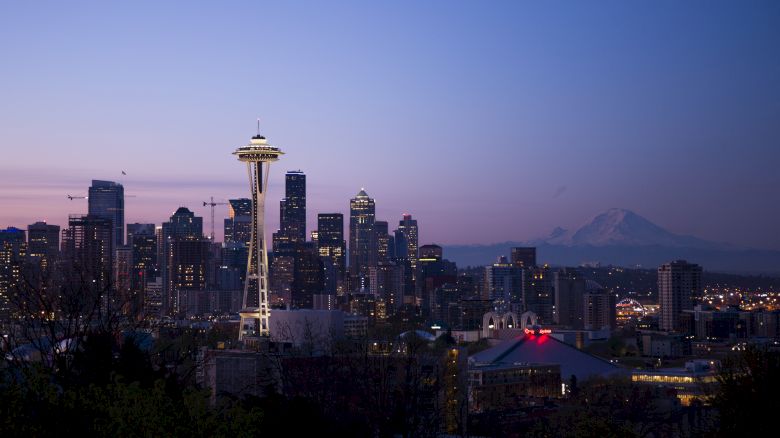 The image features a city skyline at dusk with a prominent tower in the foreground and a mountain in the background. The city lights are starting to glow.