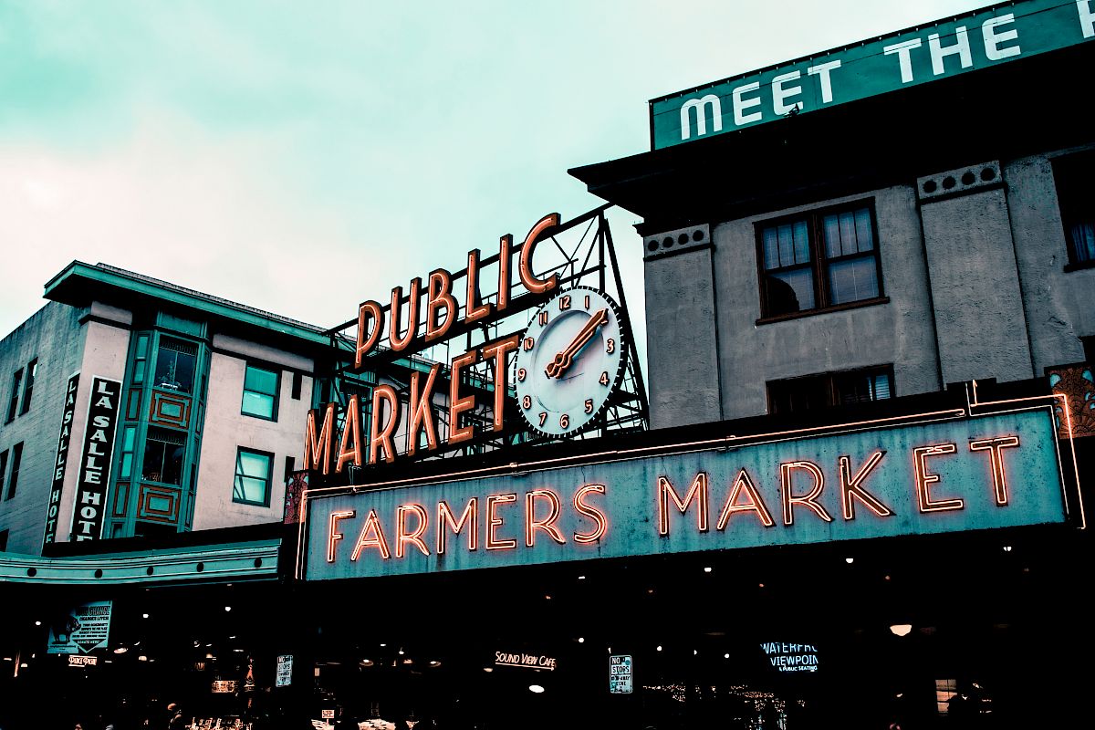 The image shows a vibrant sign for a "Public Market" and "Farmers Market" with a clock, located in a historic district with surrounding buildings.