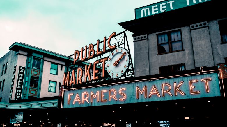 The image shows a vibrant sign for a "Public Market" and "Farmers Market" with a clock, located in a historic district with surrounding buildings.