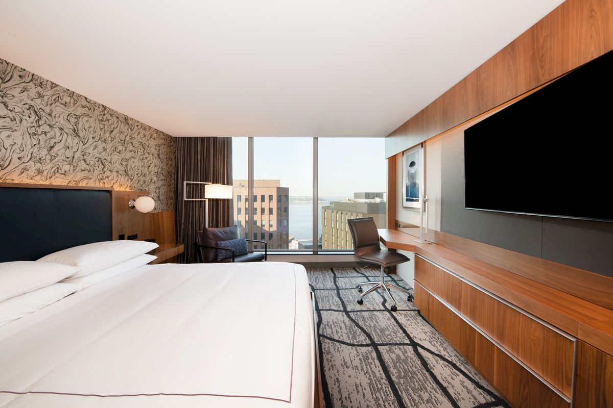 A modern hotel room features a large bed, flat-screen TV, desk, chair, and a window offering a city view in the background.