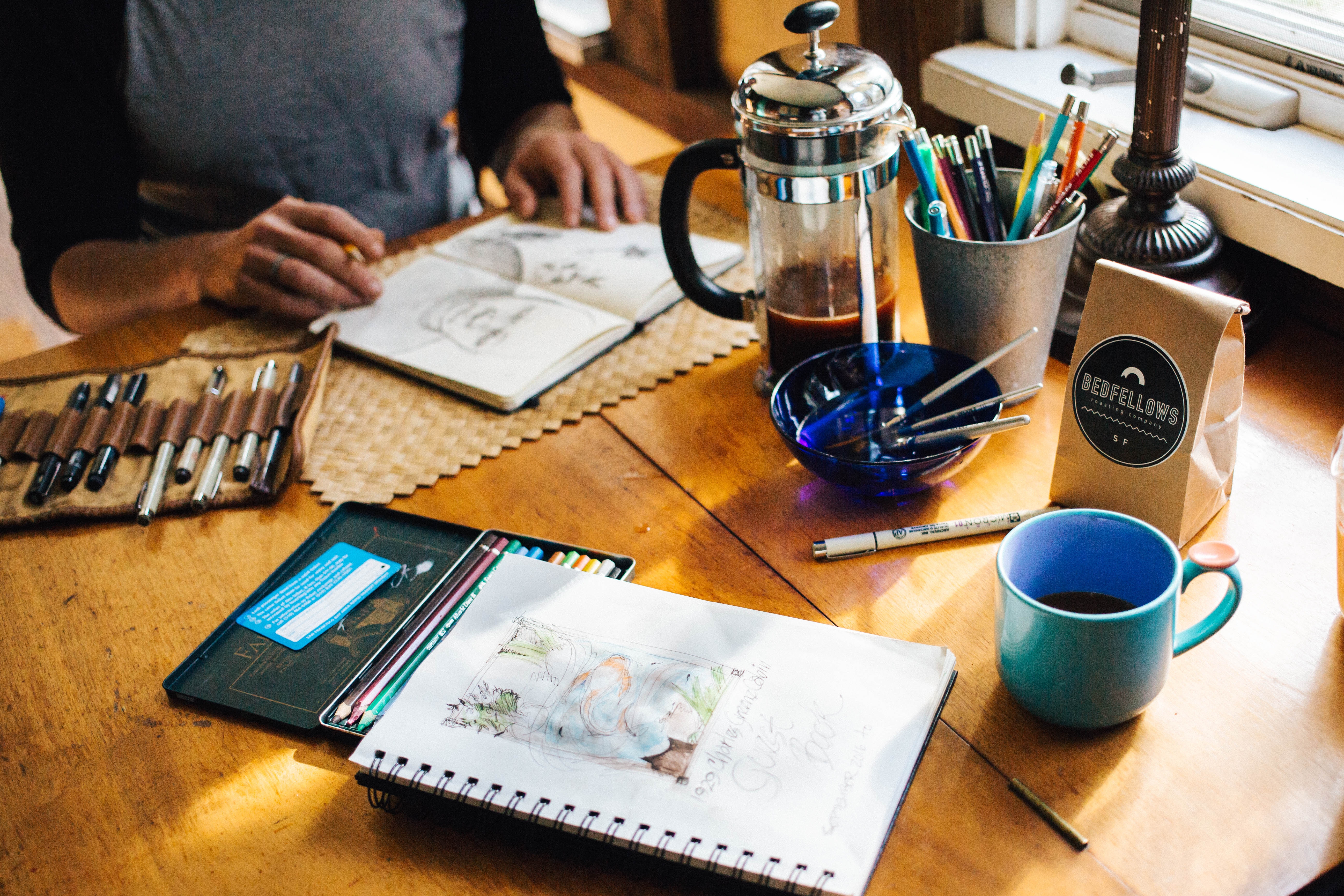 A person is sketching at a wooden table with art supplies, a coffee press, a cup of coffee, and a bag of coffee beans nearby.